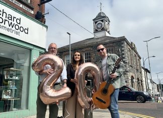 Three individuals standing in front of a stone building with a clock tower, holding balloons shaped as the numbers 20. One person also holds a guitar.