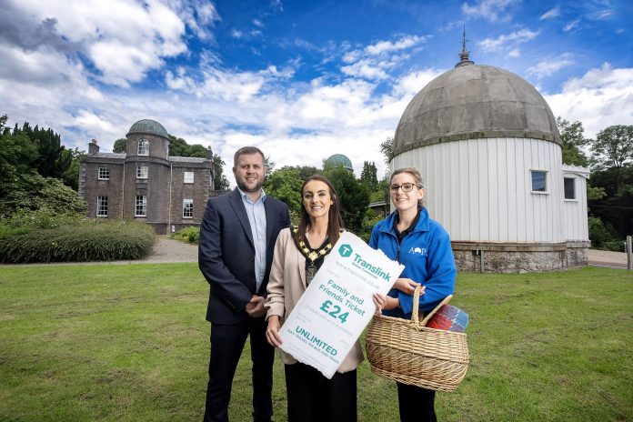 Three people standing outside an observatory with a large telescope dome. One individual holds a sign and another holds a picnic basket. The background features lush greenery and the observatory building.