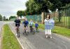 Four children on bicycles smile as they ride along a paved road next to a grassy area, accompanied by an adult holding a sign reading "Small Settlements Regeneration Programme." Trees and a green fence line the background.