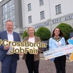 Four individuals standing in front of the Armagh City Hotel holding signs related to the "#CrossBorderJobFair Labour Market Partnership Working Together" event.