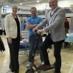 Three adults at an indoor event, one of whom is riding an electric scooter, while the other two stand beside them, smiling. A banner reading 'PostSmart' is visible in the background.