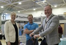 Three adults at an indoor event, one of whom is riding an electric scooter, while the other two stand beside them, smiling. A banner reading 'PostSmart' is visible in the background.