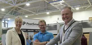 Three adults at an indoor event, one of whom is riding an electric scooter, while the other two stand beside them, smiling. A banner reading 'PostSmart' is visible in the background.