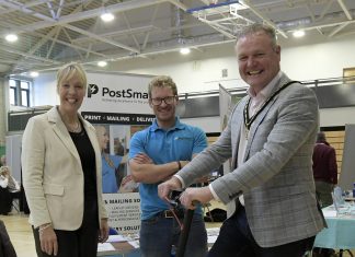 Three adults at an indoor event, one of whom is riding an electric scooter, while the other two stand beside them, smiling. A banner reading 'PostSmart' is visible in the background.