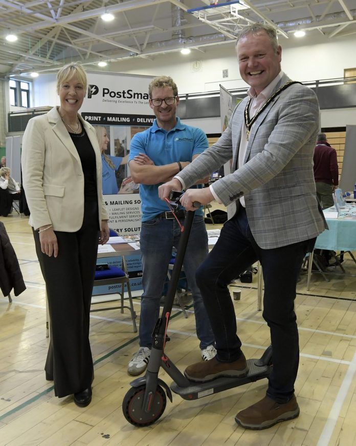 Three adults at an indoor event, one of whom is riding an electric scooter, while the other two stand beside them, smiling. A banner reading 'PostSmart' is visible in the background.