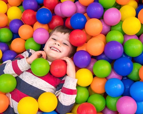 A child in lying in a soft play area surrounded by colourful small balls.