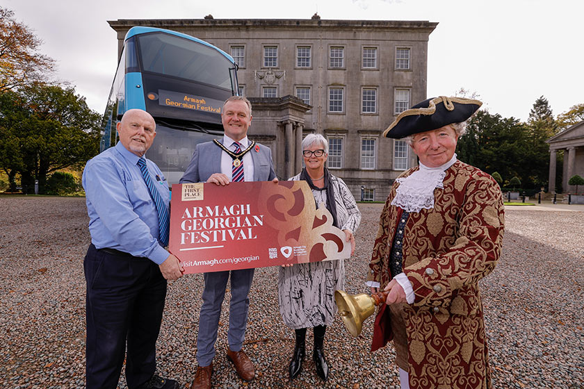 Four people standing in front of a Translink bus, parked in front of the Palace Building Armagh.